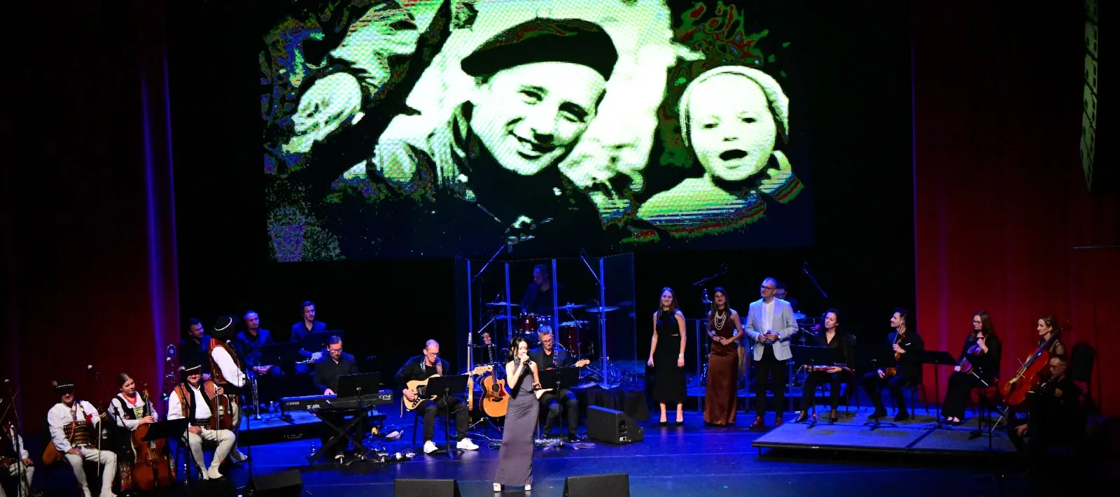 Singer and musicians, with a folk band in Highlander costumes, performing on stage under a projected archival photo