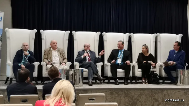 Six panelists on stage during a discussion session, one gesturing as he speaks into a microphone