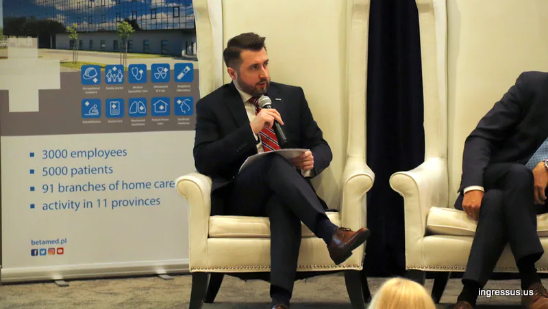 Panelist in a white armchair speaking into a microphone next to a BetaMed roll-up banner