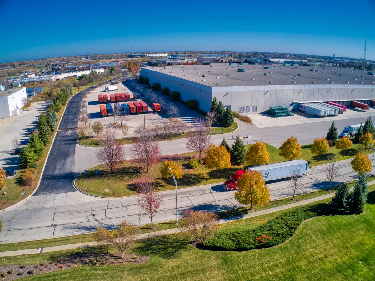 Aerial view of a red semi-truck with a white trailer driving past a large distribution warehouse on a sunny autumn day