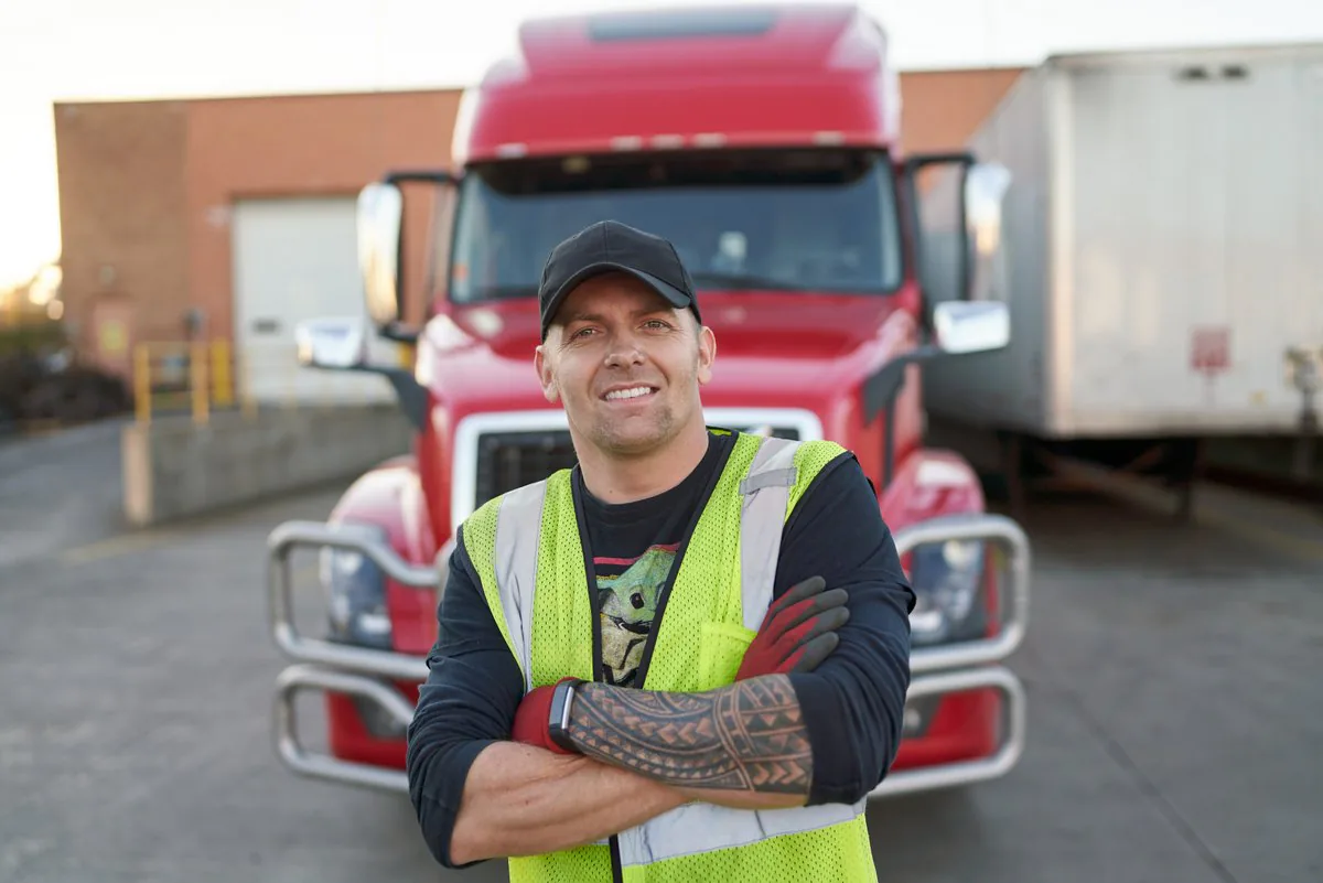 Driver in a high-visibility vest and work gloves with arms crossed in front of a red semi-truck at a loading dock