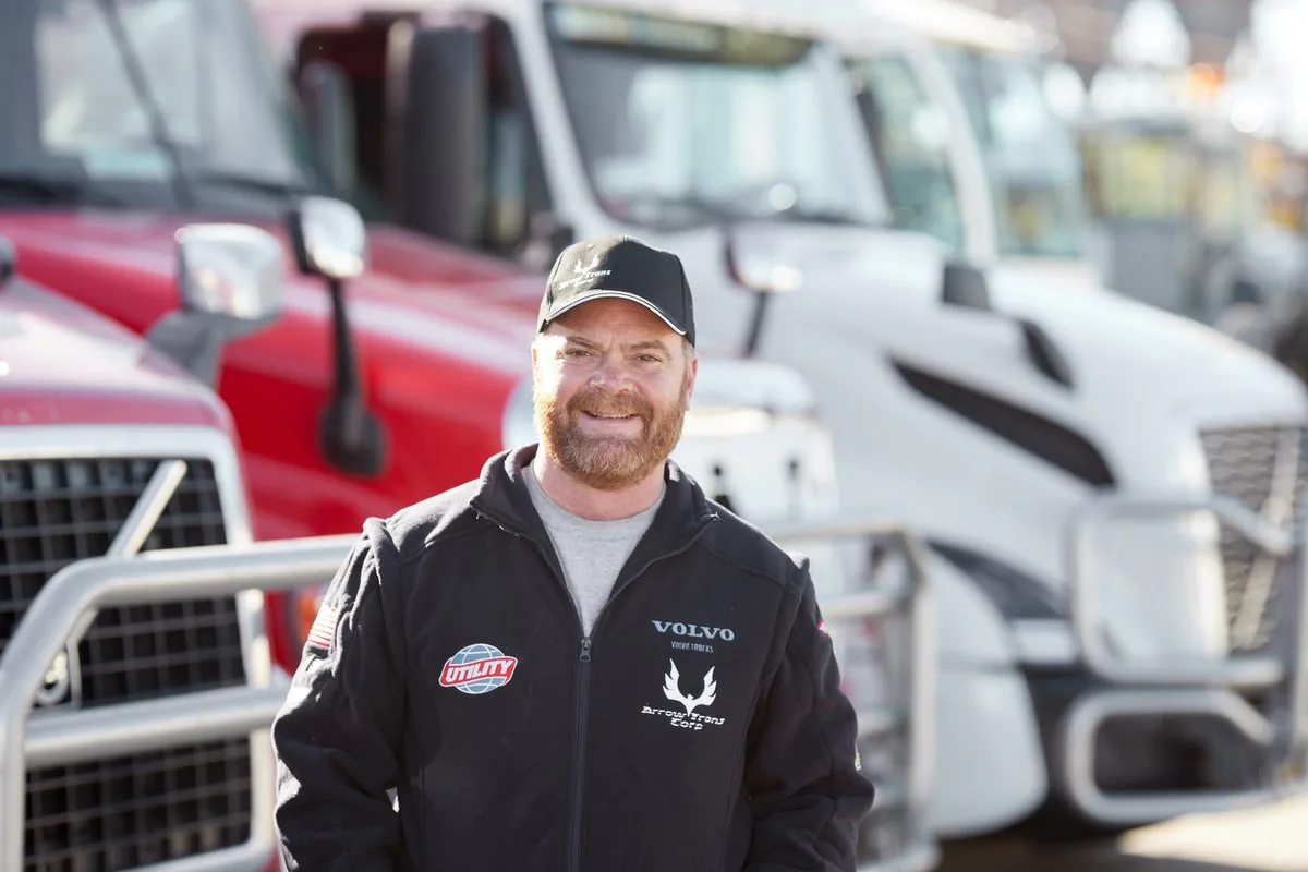 Smiling driver in a black jacket with Volvo and Arrow Trans Corp logos in front of red and white trucks