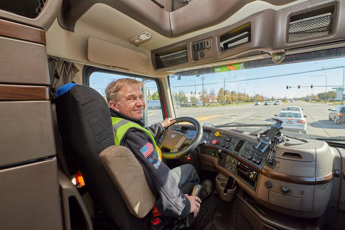 Smiling driver in a high-visibility vest at the wheel of a Volvo truck, with the road and traffic lights ahead
