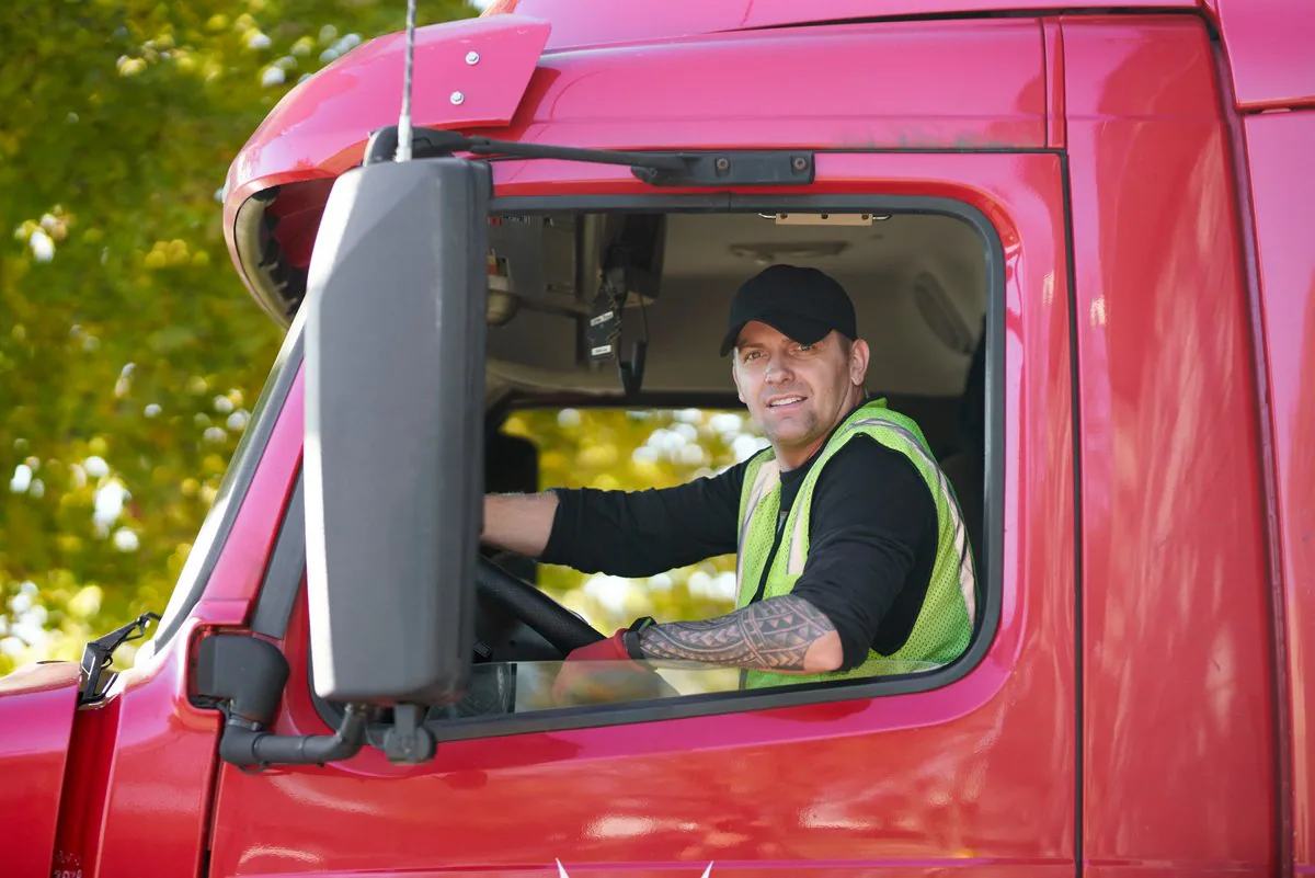 Smiling driver in a cap and high-visibility vest looking out of the window of a red truck cab