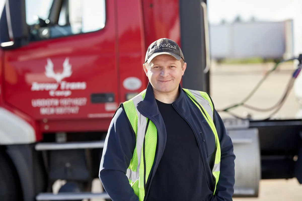 Smiling driver in a high-visibility vest and cap standing in front of a red Arrow Trans Corp truck