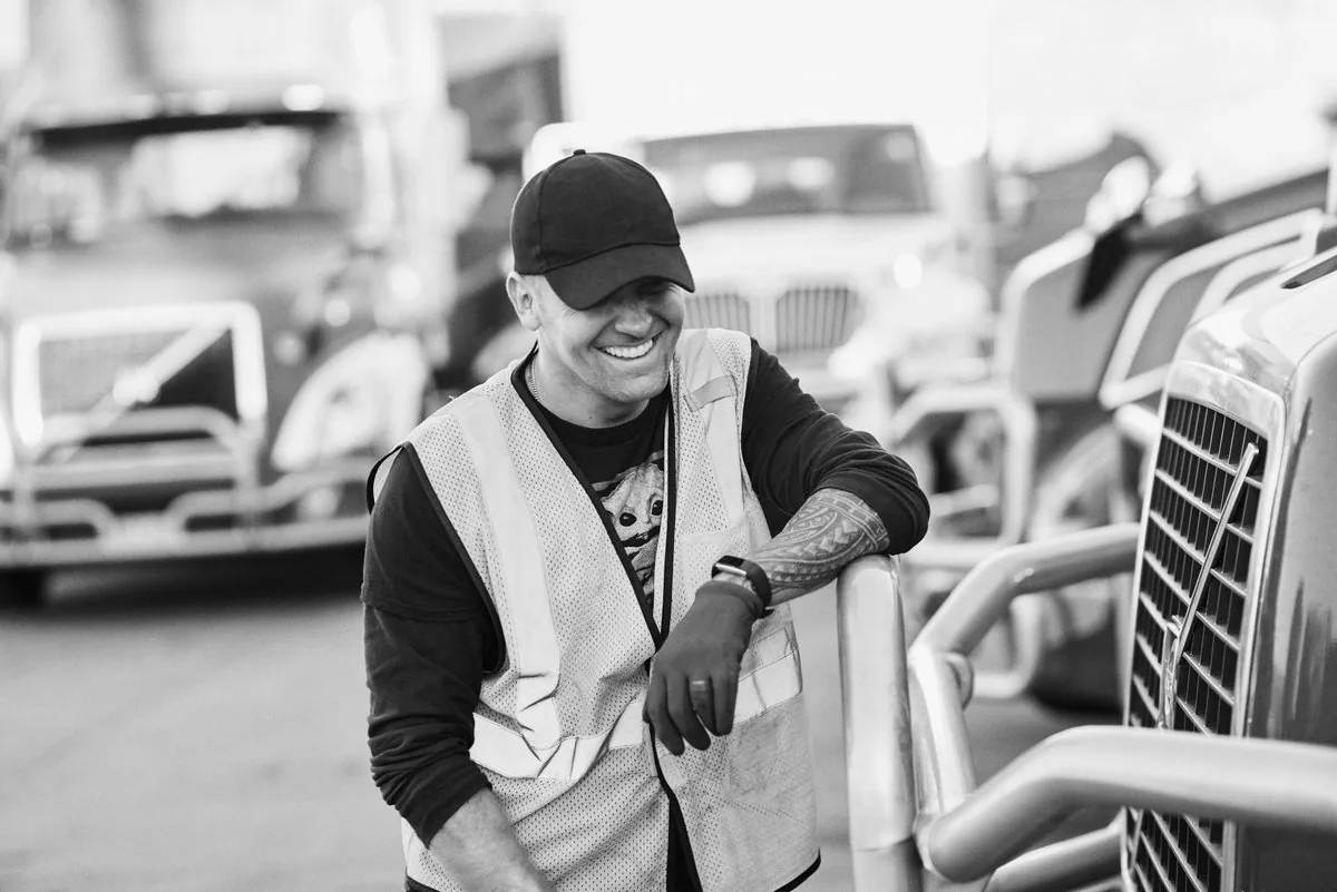 Black and white photo of a laughing driver in a safety vest leaning on a truck's chrome grille guard