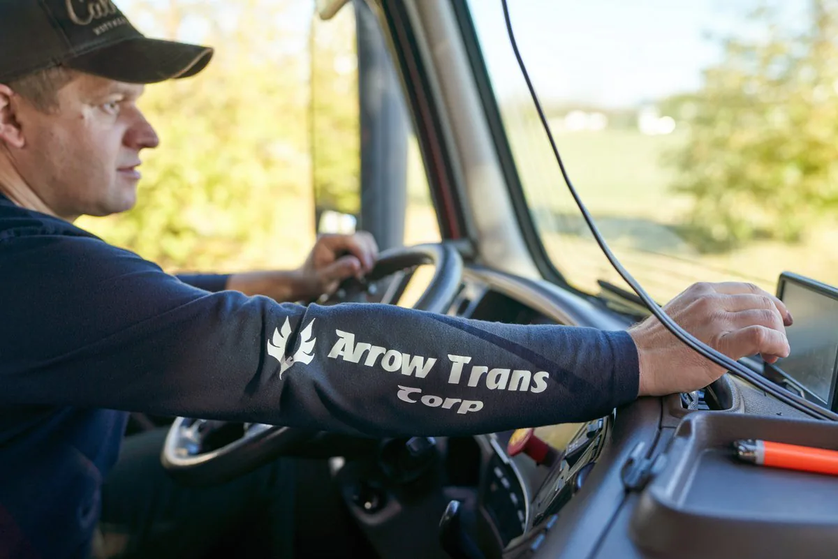 Driver at the wheel of a truck, his sleeve printed with the Arrow Trans Corp logo, reaching for the GPS screen