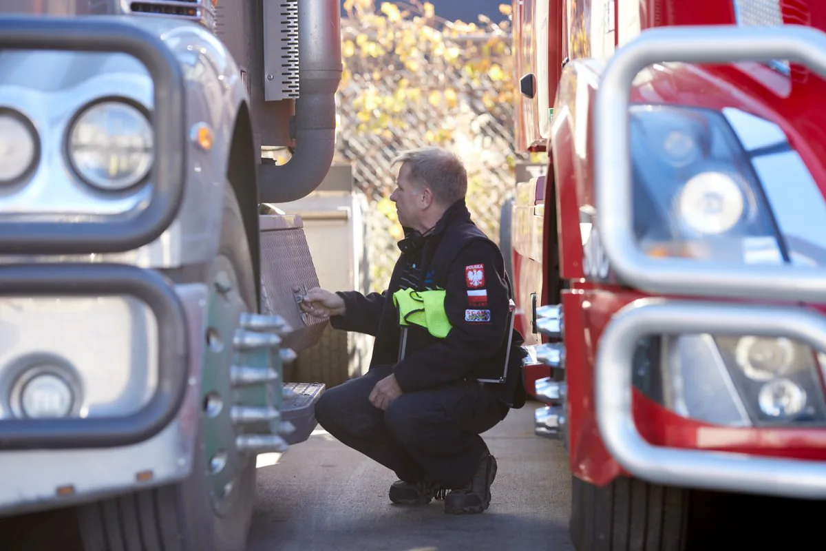 Driver crouching between two trucks during a pre-trip inspection, a Polish flag patch on his sleeve