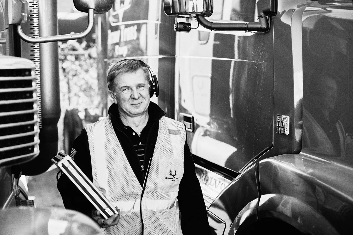 Black and white portrait of a driver in an Arrow Trans Corp safety vest with a headset and clipboard between two trucks