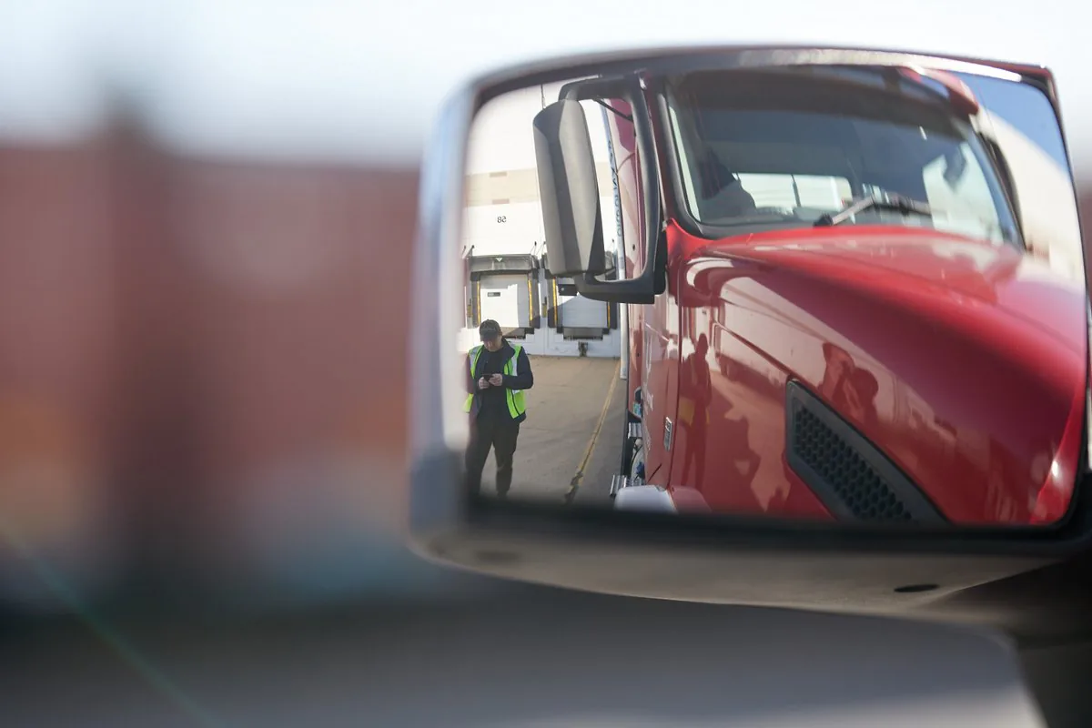 Truck side mirror reflecting a red cab and a driver in a high-visibility vest checking his phone at the loading docks