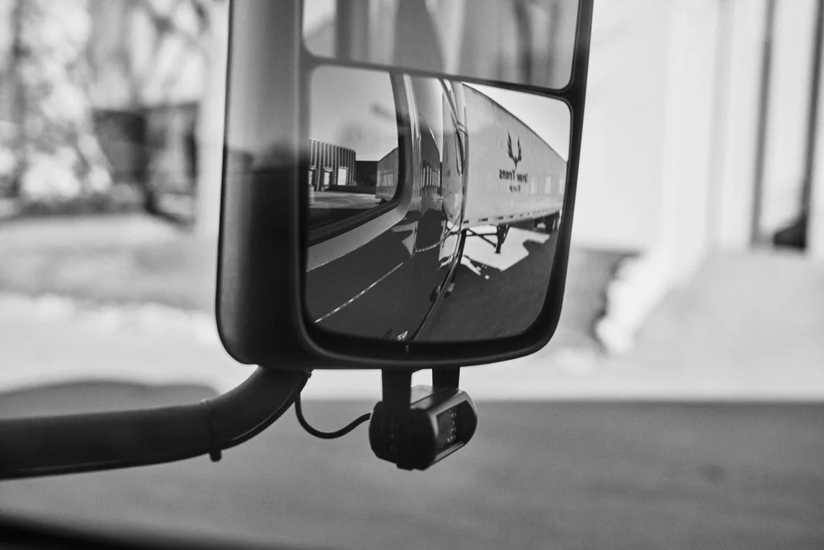 Black and white view of a truck side mirror reflecting a white Arrow Trans Corp trailer at a warehouse