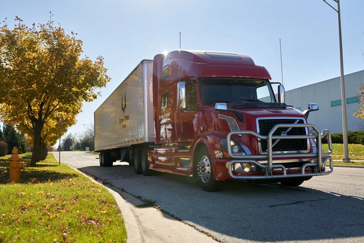 Red Volvo semi-truck pulling a white Arrow Trans Corp trailer, parked on a street lined with autumn trees