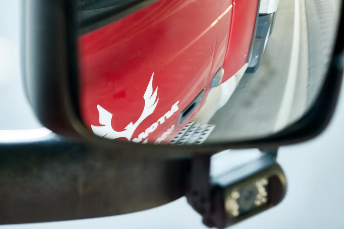 Truck side mirror reflecting a red Arrow Trans Corp cab with the company's white logo on the door