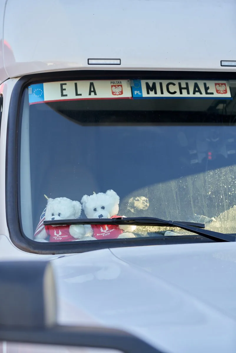 Two white teddy bears in red Arrow Trans Corp shirts behind a truck windshield decorated with Polish-style name plates