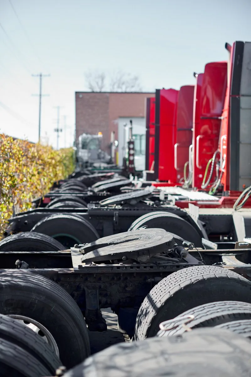 Row of red truck tractors seen from behind, with fifth wheel couplings and tires lined up in the yard