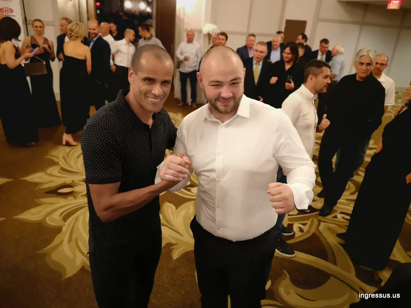 Two men posing with clasped hands and a raised fist among guests in the ballroom at the Soccer Legends Gala
