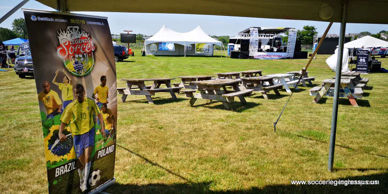 Soccer Legends Cup roll-up banner under a tent at a picnic ground with picnic tables and an outdoor stage