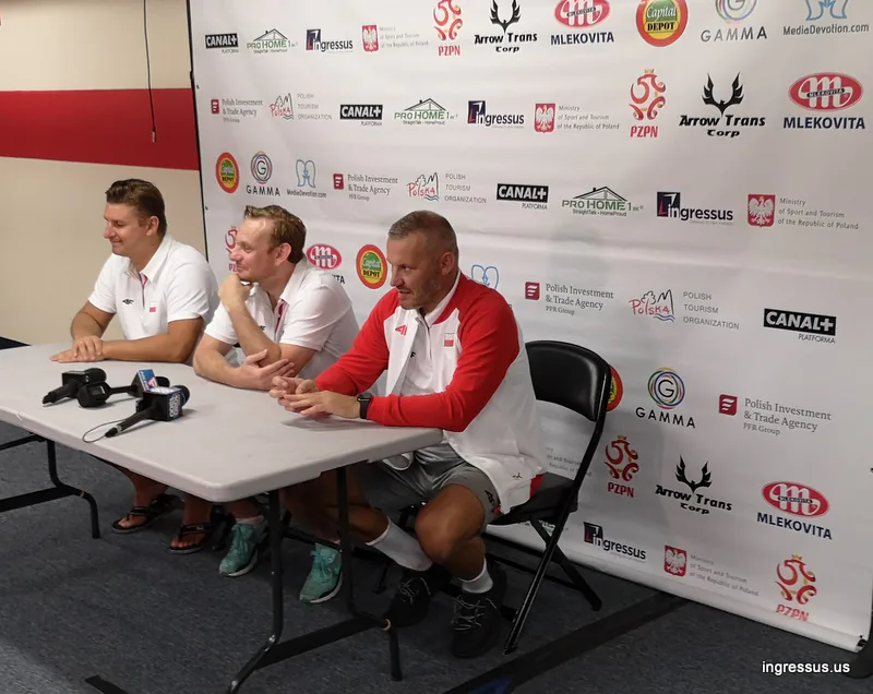 Three men in Poland team apparel at a press conference table with microphones in front of the Soccer Legends Cup sponsor wall
