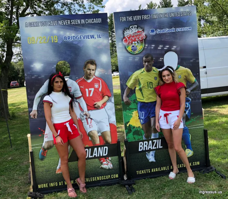 Two promoters in Poland and Brazil themed outfits posing at the Soccer Legends Cup photo boards at an outdoor event
