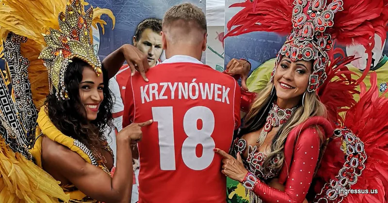 Two samba dancers in feathered costumes pointing at the back of a Poland jersey with Krzynówek 18 printed on it
