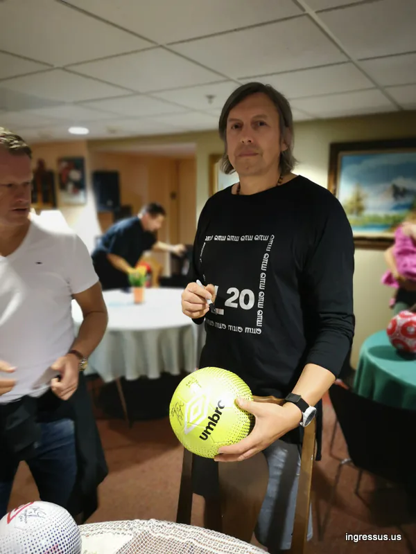 Man signing a yellow Umbro soccer ball at an indoor gathering ahead of the Soccer Legends Cup