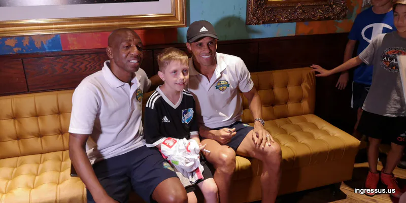 Two men in Soccer Legends Cup polo shirts posing on a sofa with a young fan holding a signed jersey at a meet and greet