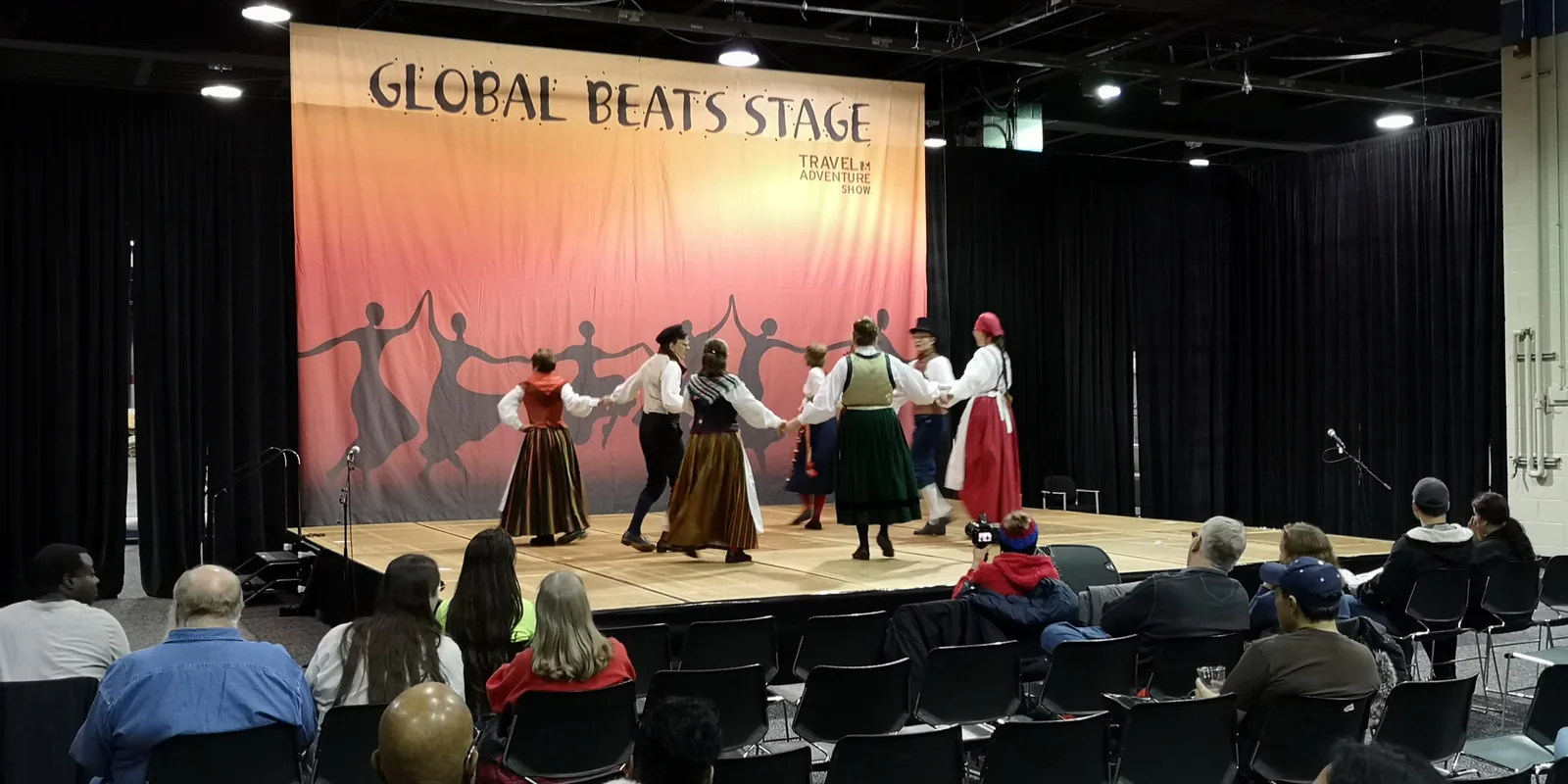 Folk dancers in traditional costumes performing on the Global Beats Stage for a seated audience at the Chicago Travel & Adventure Show 2019