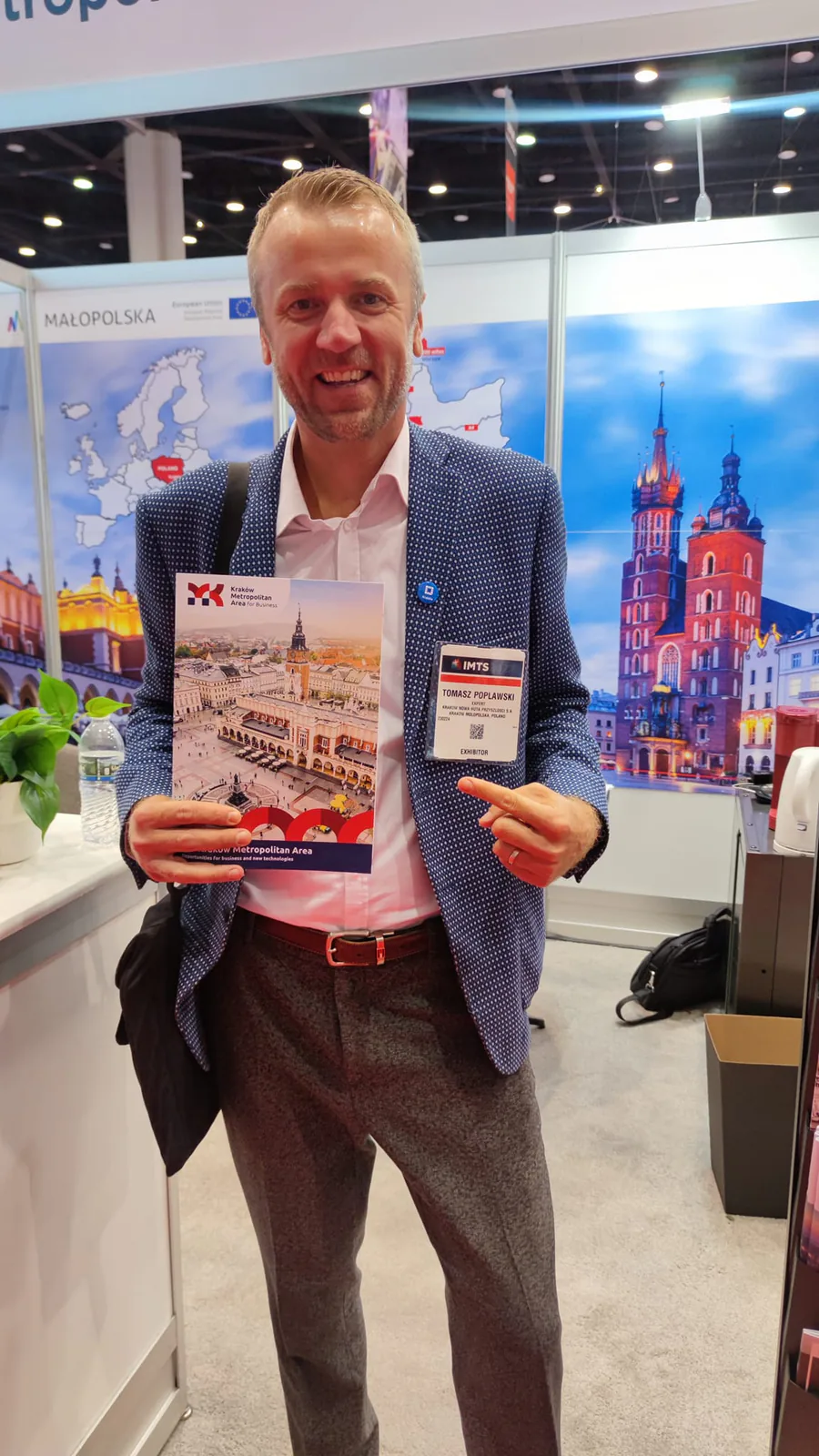 Tomasz Popławski, wearing his IMTS exhibitor badge, holding a Kraków Metropolitan Area brochure at the booth