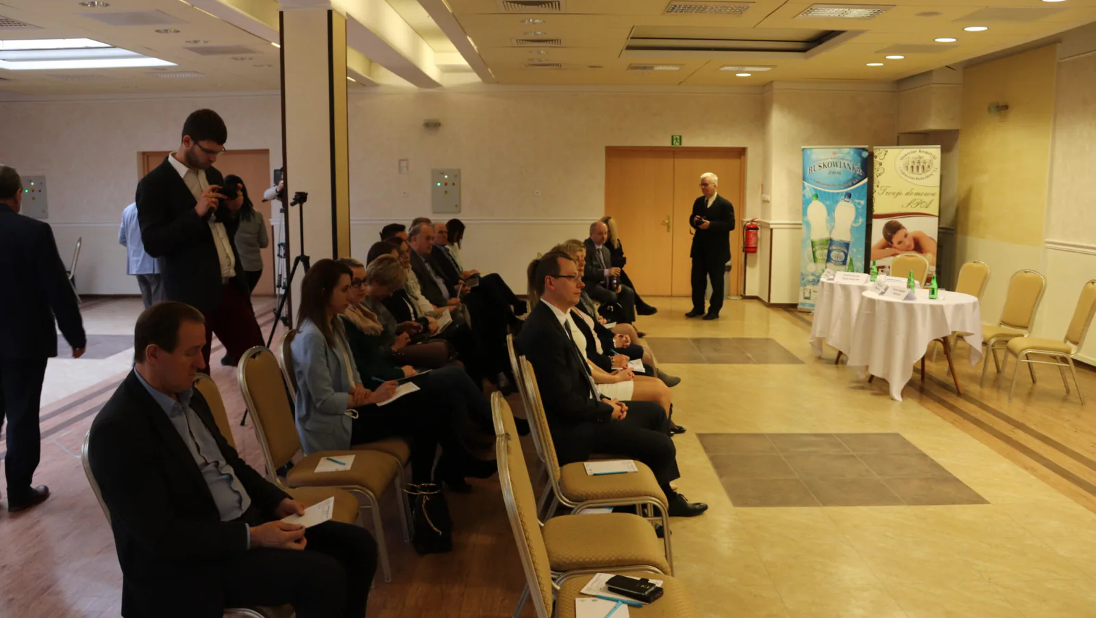 Guests seated at the Busko-Zdrój seminar, with photographers, a camera on a tripod and the speakers' table in the background