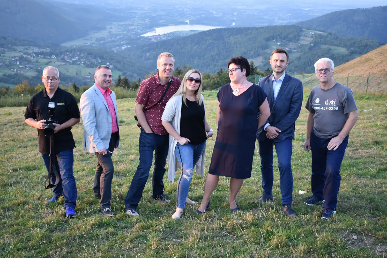 Seven members of the press mission posing on Żar mountain, with a valley and a lake below them