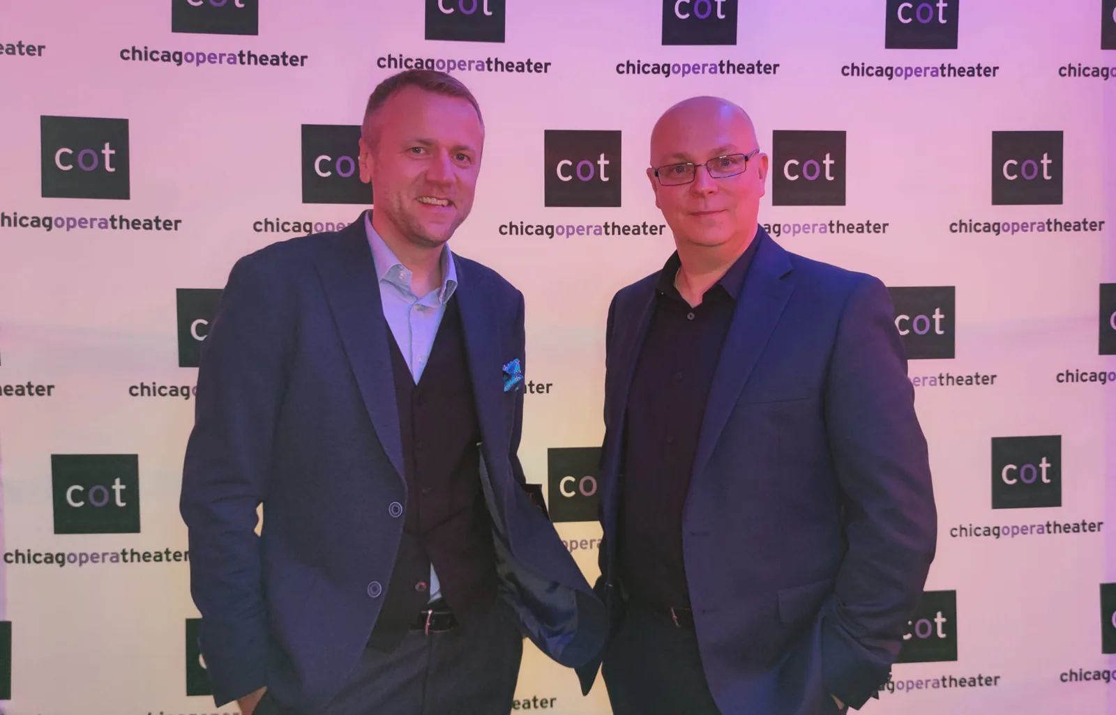 Two men in blazers posing in front of the Chicago Opera Theater logo backdrop at the King Roger event