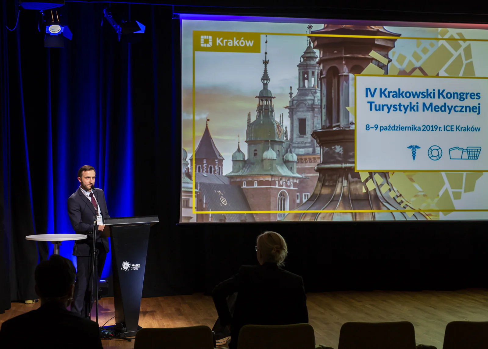 Speaker at a Kraków Congress Centre lectern next to the congress title screen with Wawel Castle towers