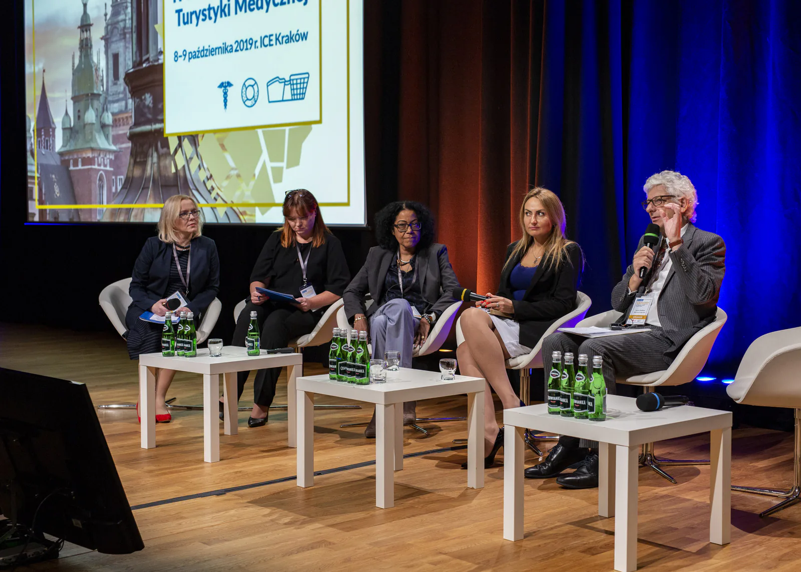 Five panelists at white tables on stage, one speaking into a microphone, during a medical tourism congress panel