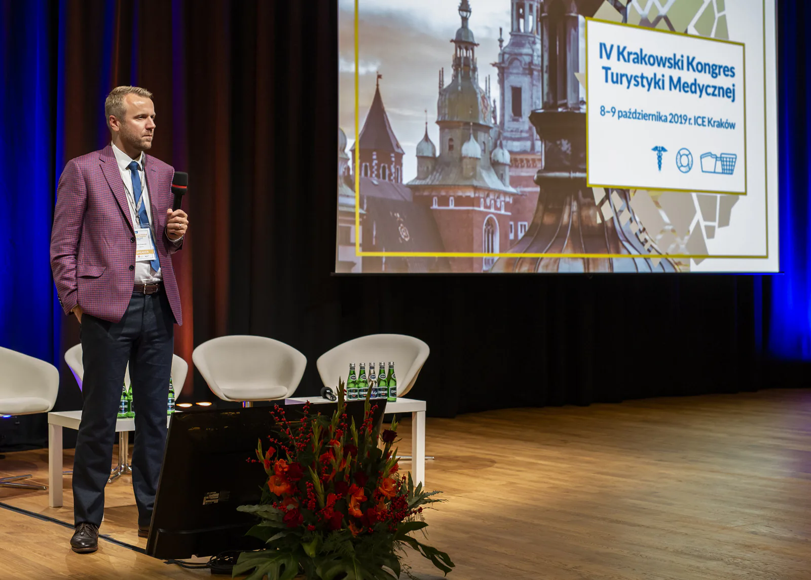 Speaker holding a microphone on stage next to flowers and the 4th Kraków Medical Tourism Congress title screen
