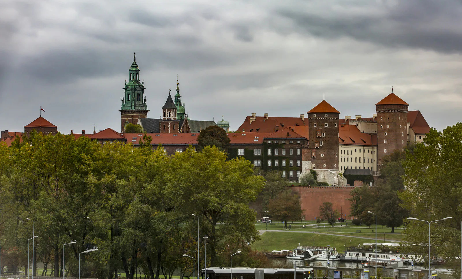 Wawel Royal Castle and Cathedral in Kraków under a cloudy autumn sky, seen across the Vistula riverbank