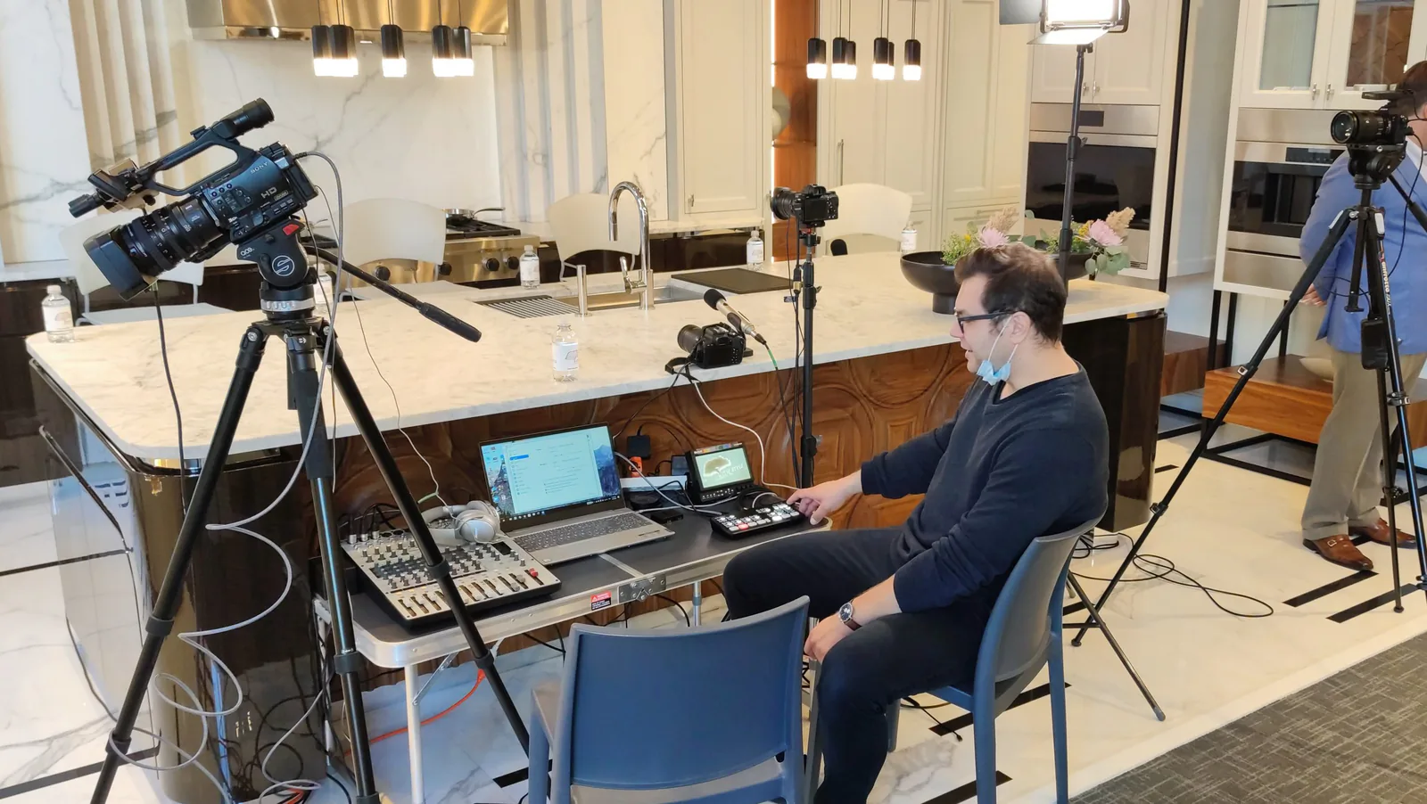 Video operator in a face mask at the streaming desk, with cameras on tripods around a kitchen showroom set