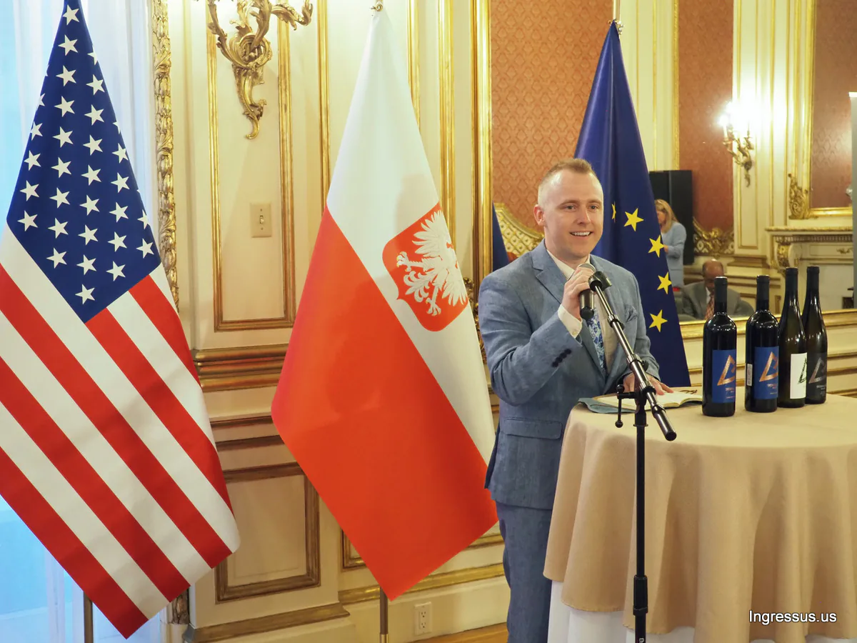 Speaker with a microphone beside a table of wine bottles, in front of U.S., Polish and EU flags, during the Małopolska event