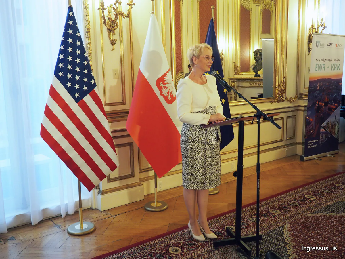 Speaker at a lectern beside U.S., Polish and EU flags and a New York/Newark to Kraków EWR to KRK flight banner
