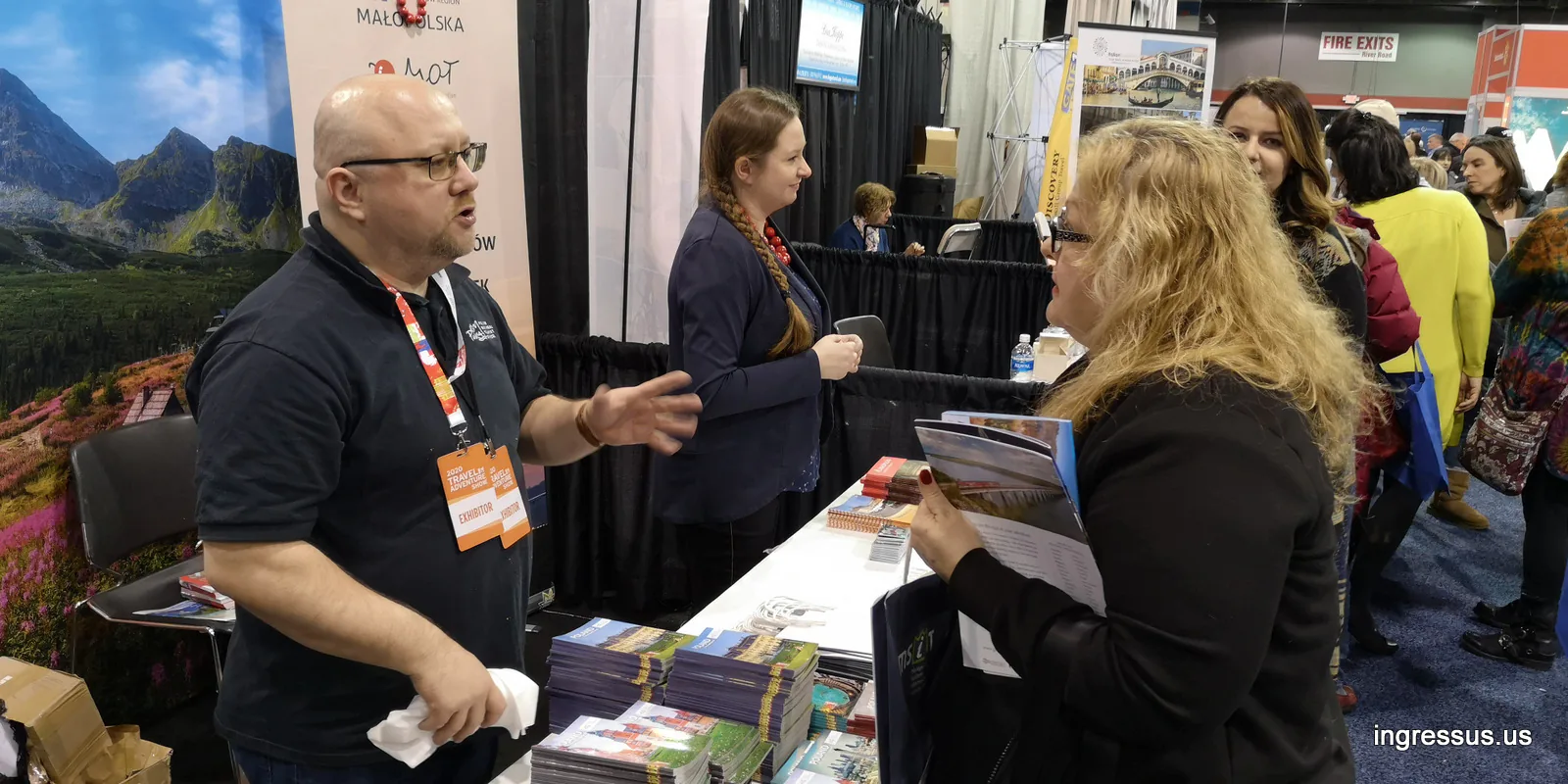 Exhibitor with a Travel & Adventure Show badge talking with a visitor holding a Małopolska brochure at the booth