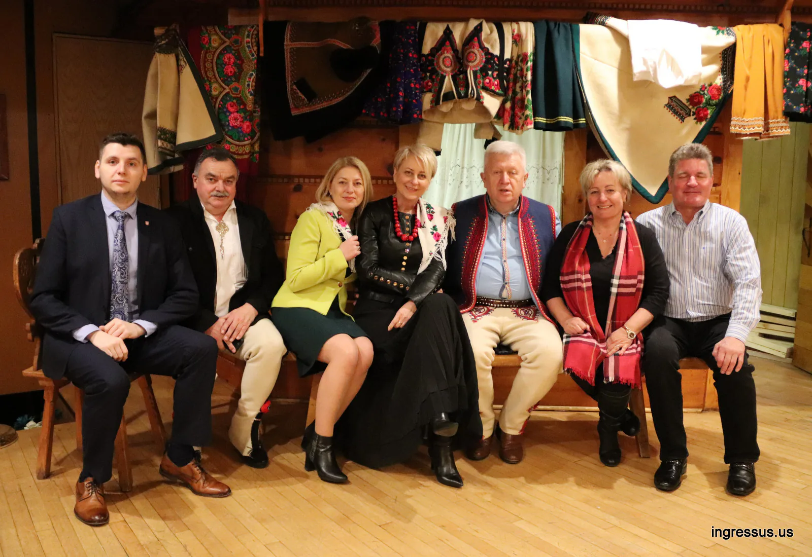 Group seated on a wooden bench in a hall decorated with Polish highlander folk costumes and embroidered garments