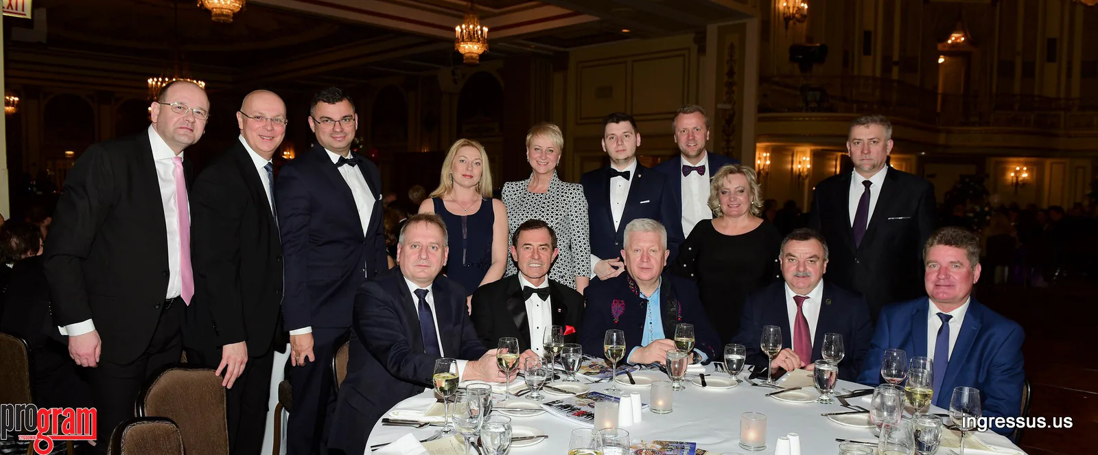 Guests in formal evening wear posing around a banquet table in a chandeliered ballroom at a gala dinner