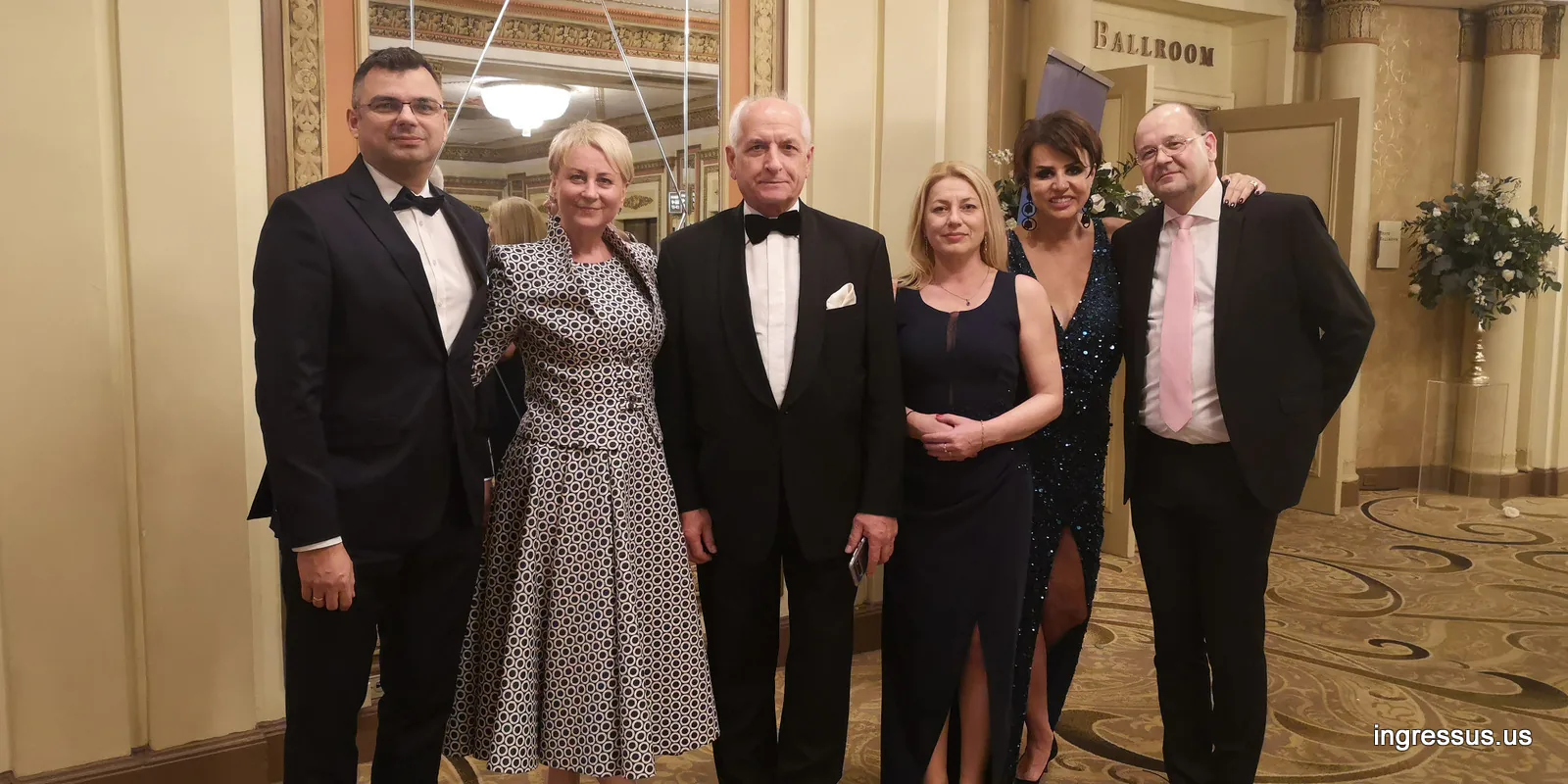 Six guests in formal evening wear posing by a hotel ballroom entrance on a gala night