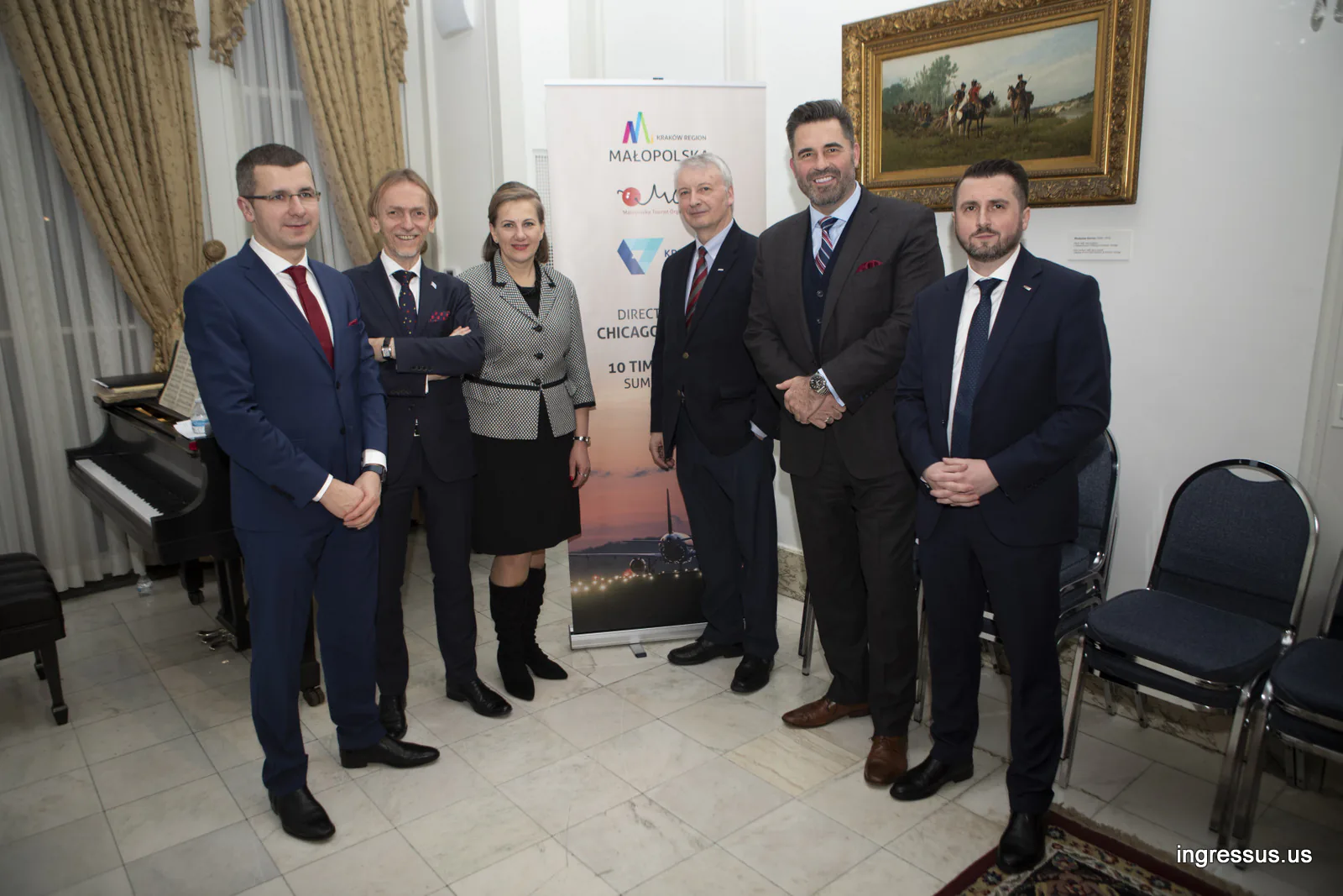Six guests posing by a Małopolska and Kraków Airport direct flights banner in a consulate room with a grand piano and a painting