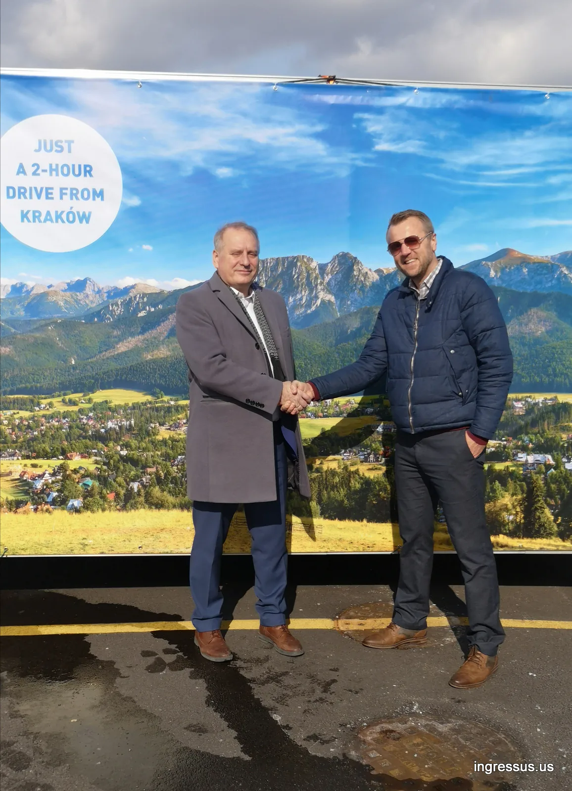 Two men shaking hands in front of an outdoor banner with mountain scenery and the words Just a 2-hour drive from Kraków