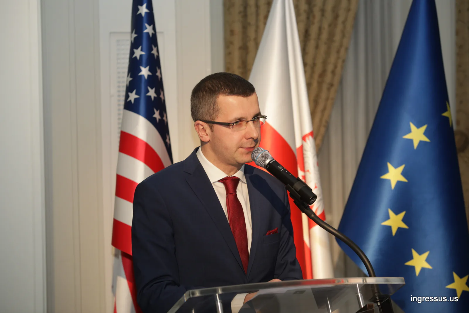 Speaker in a red tie addressing guests from the podium, with U.S., Polish and EU flags behind him, at the Polish Consulate General
