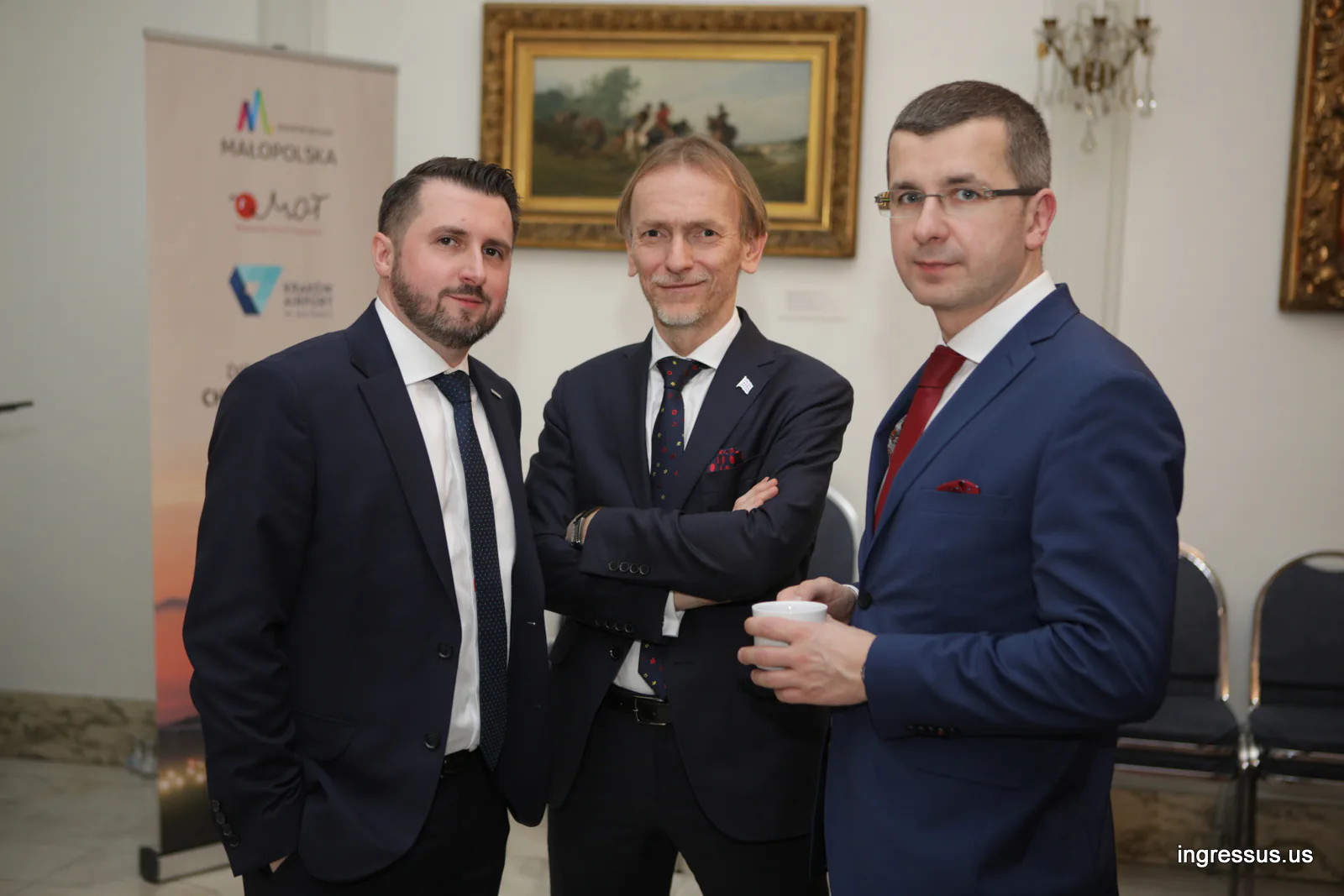 Three men in suits talking by a Małopolska, MOT and Kraków Airport banner at the Polish Consulate General reception