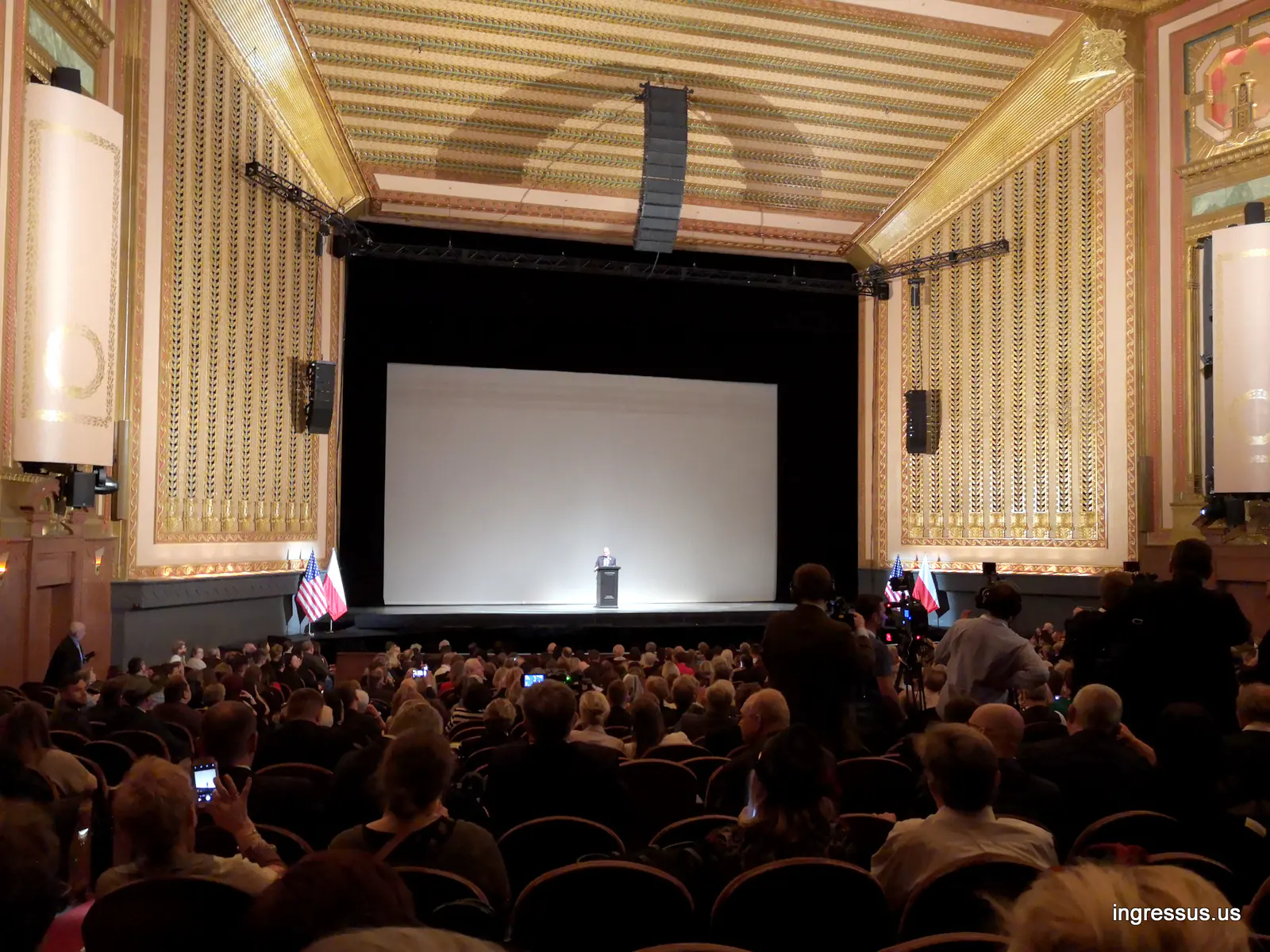 Speaker at the podium on the Lyric Opera House stage before the Poland: The Royal Tour screening, with a full audience