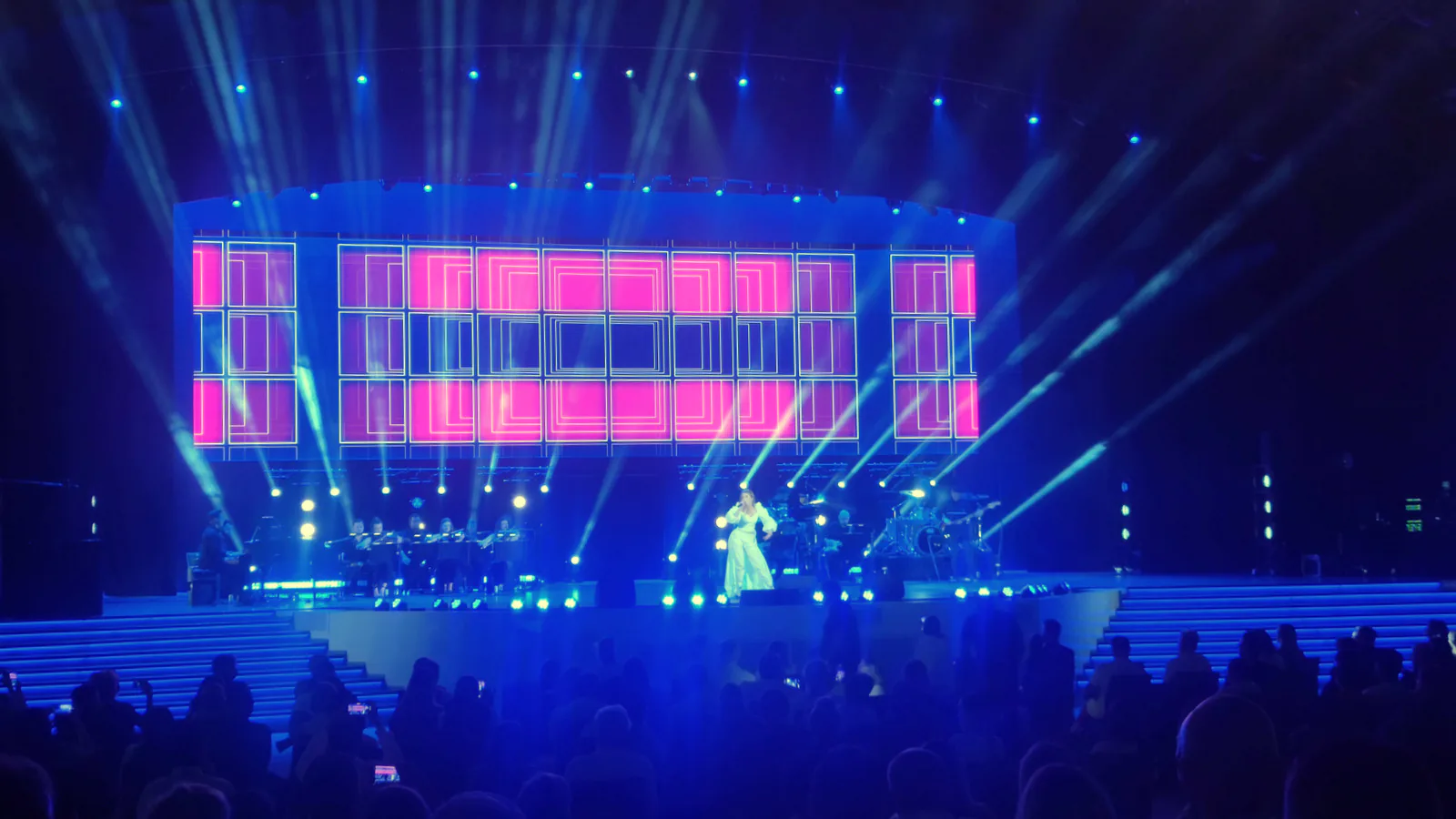 Singer in a white gown performing with a band on a stage lit by blue beams and pink screens during an evening concert