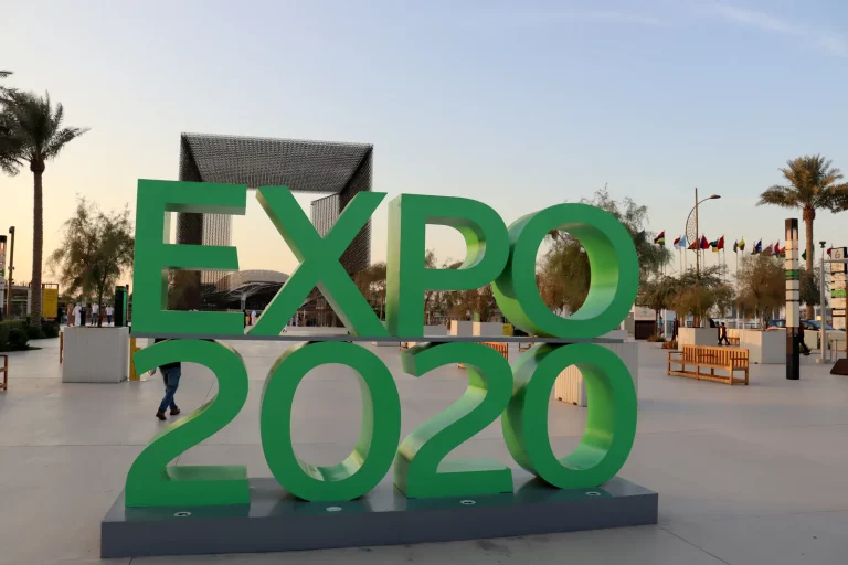 Green EXPO 2020 letters at the Expo 2020 Dubai site, with a gateway structure, palm trees and national flags behind