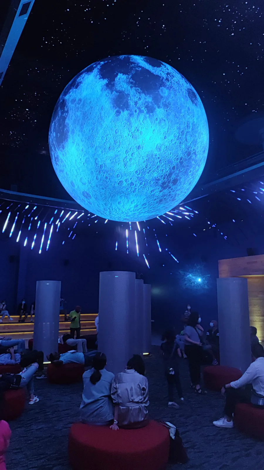 Visitors seated beneath a giant glowing moon globe in a dark, star-lit hall at Expo 2020 Dubai